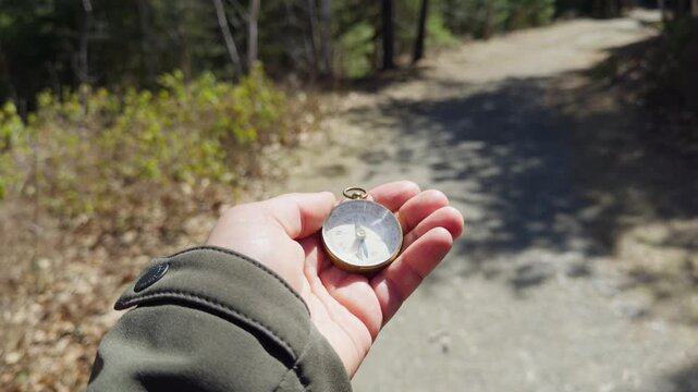A hand holds a vintage compass as it points toward a winding trail. Surrounded by lush trees and sunlight, the journey unfolds into the beauty of the great outdoors