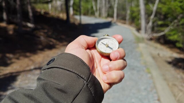 A person holds a compass while standing on a peaceful forest path. Lush trees surround the trail, hinting at an adventure in the great outdoors. Sunlight filters through the leaves above
