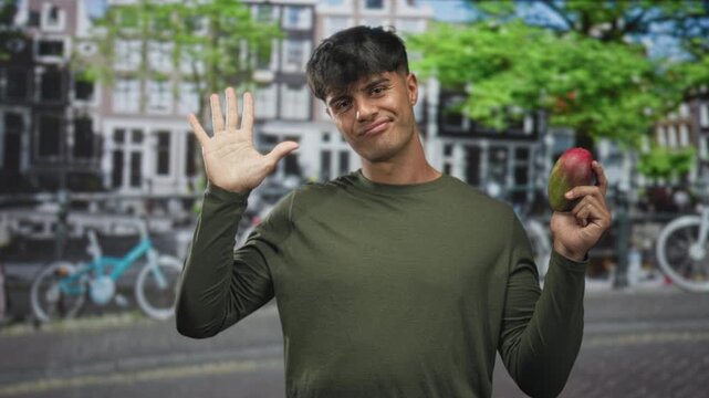 Man holding a ripe mango and waving with raised hand on street beside bicycles and canal buildings, wearing olive green shirt and smirking; playful greeting.