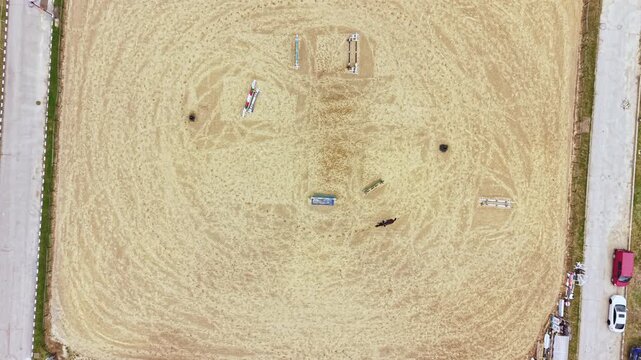 A sandy arena shows horse training with several jumps and obstacles arranged in different positions. Two horses are practicing while a vehicle is parked nearby.