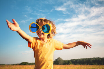 A happy little boy wearing large, funny sunglasses enjoys the sun. The boy stands against a blue sky on a sunny day © vvvita