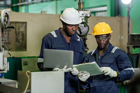 Two African technicians stand with laptop and clipboard near industrial machine, checking document detail and digital record for maintenance review, production tracking, and factory coordination.