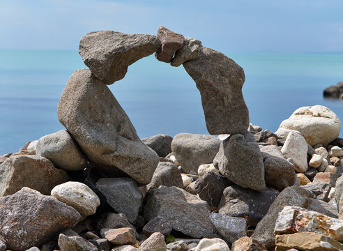 Stone sculpture formed by balancing rocks on a beach, with calm sea and blue sky in the background creating a serene coastal scene