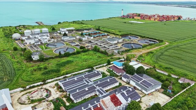 Aerial coastal treatment plant with solar arrays, circular settling ponds, lagoon basins, access roads, maintenance buildings, surrounding mangrove buffer,
