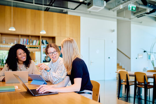Three diverse women working together on laptops and a tablet, discussing ideas in a comfortable coworking environment