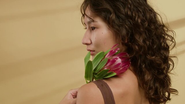 Curly-haired girl with no makeup turning slowly to camera, with pink protea flower on her left shoulder