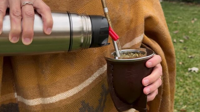 Close-up of an older woman's hands in a yellow poncho pouring steaming water to prepare a traditional yerba mate infusion in a leather cup with a bombilla metal straw. Autumn background