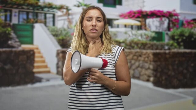 Woman holding a megaphone with hand on neck while looking upward on street by white building and stone wall; vocal strain fatigue.