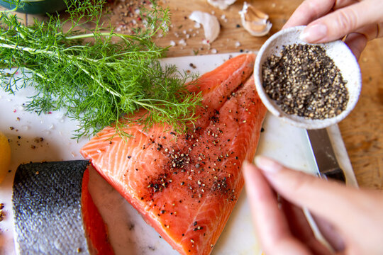 Salmon fillet being seasoned with pepper, garnished with fresh dill.