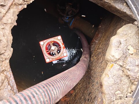 View looking down into a flooded utility manhole with a thick suction hose and a red metal water valve during emergency pipeline maintenance
