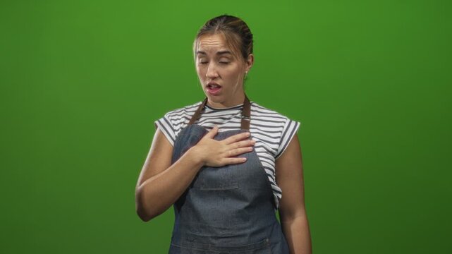 Woman in denim apron with hand on chest touching and uneasy grimace, striped shirt visible in studio; concern apology.