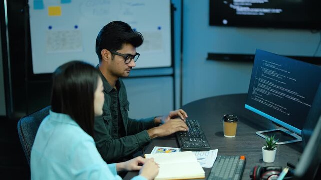 Two diverse software developers collaborate on coding at a modern office desk. A female programmer points at the computer screen while her male colleague types. Tech teamwork and problem solving.