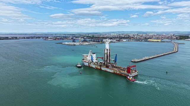 Vessel is conducting dredging operations in harbor area while a tugboat assists. City skyline is visible in the background with infrastructure along the coast.