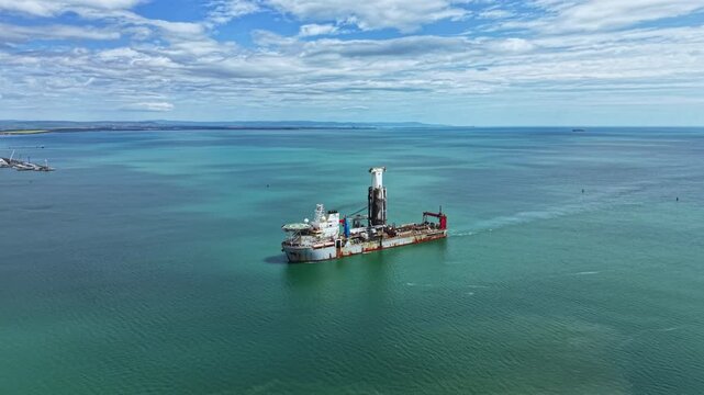 Dredging vessel moves through clear blue water, working on the seabed near a coastline under a partly cloudy sky. Background shows distant land and marine activity.