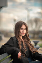 Obraz premium Young woman with long brown hair wearing black coat and blue jeans sitting on a park bench, with trees and street lamps visible in the background