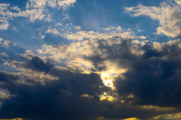 Obraz premium Bright sunbeams break through thick dark clouds in the evening sky. Contrasting golden light illuminates the edges of cumulus clouds, creating a dramatic effect.