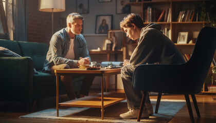 Fototapeta premium An adult man and a teenager sit in a dimly lit living room, engaged in a serious, quiet conversation across a low wooden coffee table.