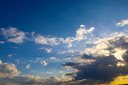Bright sunbeams break through gaps in dark cumulus clouds against a rich blue sky. Golden evening light beautifully illuminates the edges of white cloudlets, creating a picturesque contrast.