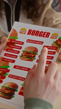 Vertical close-up of hands of unrecognizable young people studying food menu at diner, discussing burger combo offers, during friendly hangout or student gathering
