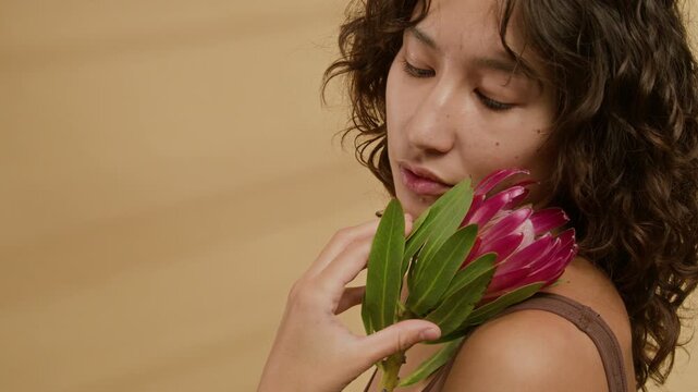 Close-up shot of young woman with curly hair gently holding protea flower near her face and looking at camera against warm-toned background