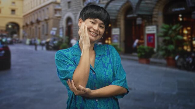 Young woman smiling with hand to cheek and bare forearm visible, standing on a cobblestone street by stone buildings and shops; joyful ease.