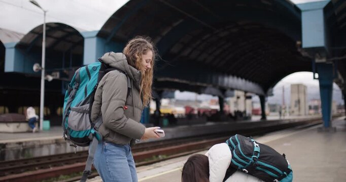 Two Young Women with Backpacks Wait at a Train Station, One Taking Photos of the Other as They Embark on a Journey Together.