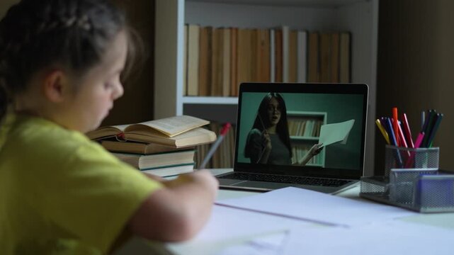 Girl studies online with teacher on laptop screen. Child writes homework during virtual lesson at home. Student learns remotely with educator. Kid does schoolwork by books and home computer.