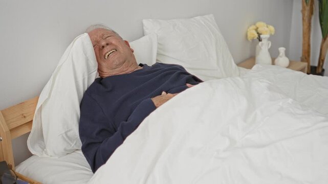 Man lying in bed mouth open hand on abdomen on wooden bedframe beside nightstand with vase and flowers in bedroom; quiet fatigue.