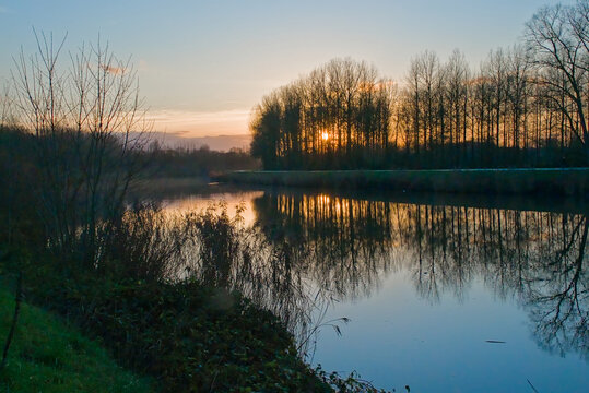 orange susnet sky over Scheldt river, with silhouettes of bare winter trees and reflections in the zqter. Ghent, Flanders, Belgium 