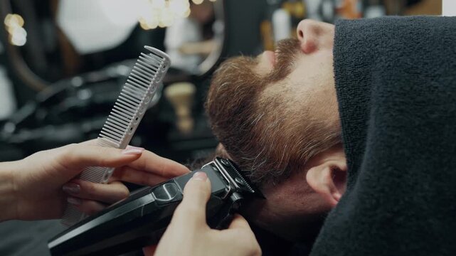 Close up of barber trimming beard with electric clipper and comb during grooming procedure in modern barbershop