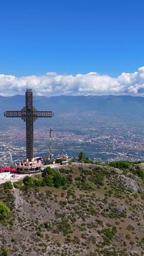 Vertical aerial drone view of the Millennium Cross on Mount Vodno in Skopje, North Macedonia. View of the large Christian monument towering above the city, surrounding hills, and urban landscape.