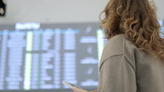 Woman watches flight board at airport, checking schedule. Curly-haired traveler stands near departures screen. She holds phone, focused on travel plans. Suitable for travel, tourism, or waiting scenes