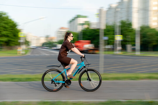 Young Caucasian Woman Cycling Urban Street On Blue Bicycle, Brown Jacket And Skirt, Brisk Morning Commute, Motion Blur Background, Trees And City