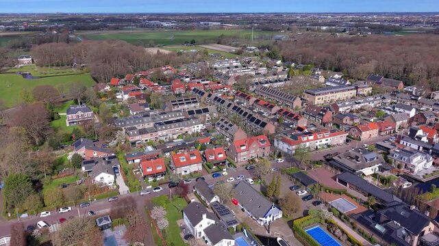 Aerial view of a charming Dutch suburban neighborhood, featuring neat rows of houses with solar panels on their roofs, blossoming spring trees, and backyard swimming pools under a bright blue sky.