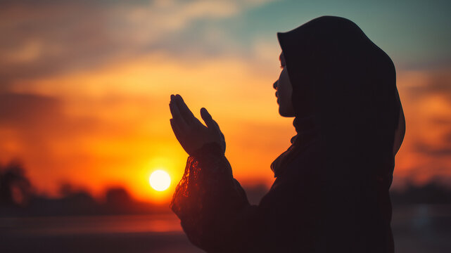 Woman in Hijab Praying at Sunset: A silhouette of a woman wearing a hijab, her hands raised in prayer, facing a vibrant sunset, conveying a sense of peace and devotion.