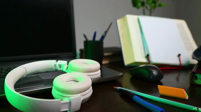 An education, school desk with a laptop, an open book, white headphones, colorful pens for art or study supplies. No people, nobody. A detail of the workspace on a wooden table. Different angles 