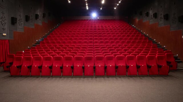 Empty cinema auditorium featuring rows of plush red velvet seats facing the screen, illuminated by overhead spotlights creating a quiet anticipatory atmosphere.