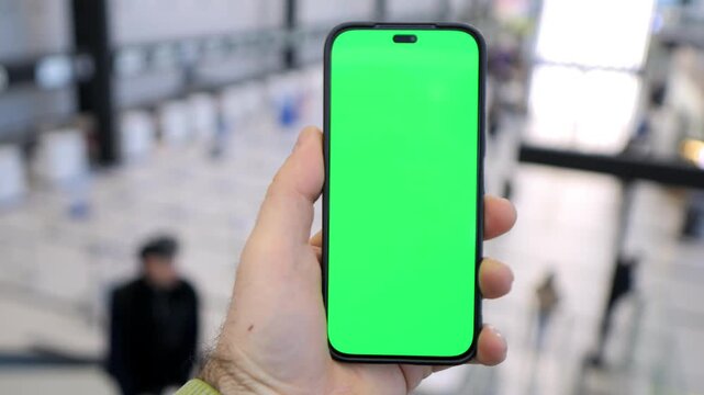 Hand holds phone with green screen. Background blurred, public space visible. Ideal for app demos, digital content, or UI mockups. Neutral expression, casual setting