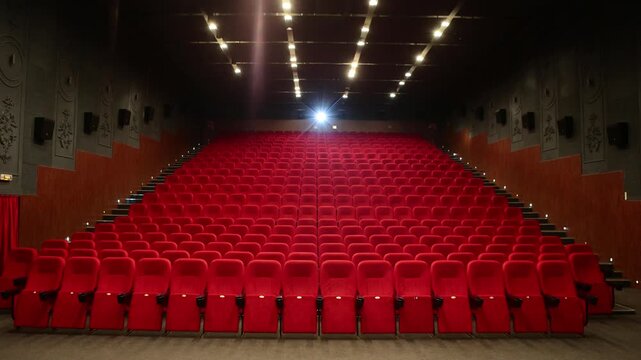 Empty cinema auditorium featuring rows of plush red velvet seats facing the screen, illuminated by overhead spotlights creating a quiet anticipatory atmosphere.