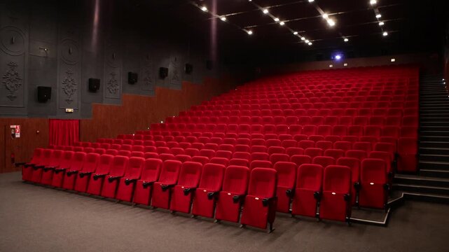 Empty cinema auditorium featuring rows of plush red velvet seats facing the screen, illuminated by overhead spotlights creating a quiet anticipatory atmosphere.