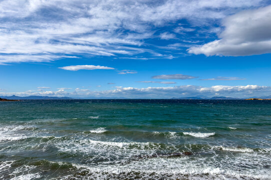 Choppy waves roll toward the shore under scattered clouds and blue sky near Vouliagmeni, Attica, Greece