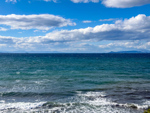 Choppy waves roll toward the shore under scattered clouds and blue sky near Vouliagmeni, Attica, Greece