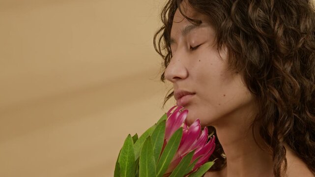 Close-up view of curly-haired girl with no makeup holding pink protea flower near her face, closing eyes