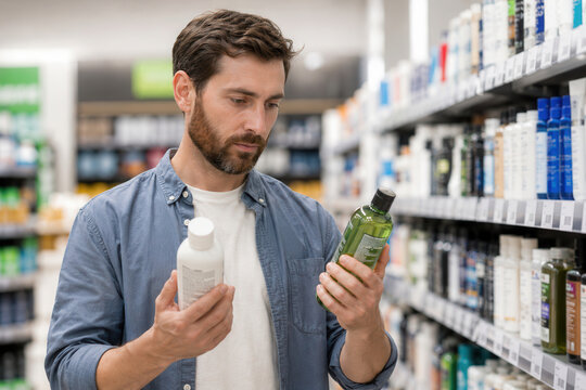 A man stands in a store aisle holding two bottles. He looks closely at the labels to compare the products. Shelves are filled with various personal care items around him.