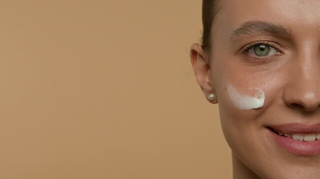Close-up half face portrait of Caucasian grey-eyed young woman applying moisturizing cream to her cheek and smiling for camera in studio