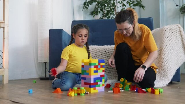 Mother and daughter playing together with colorful building block on home floor. Happy mother helps child building up tower. Family enjoys playing indoor. Daughter loves building with toy block.