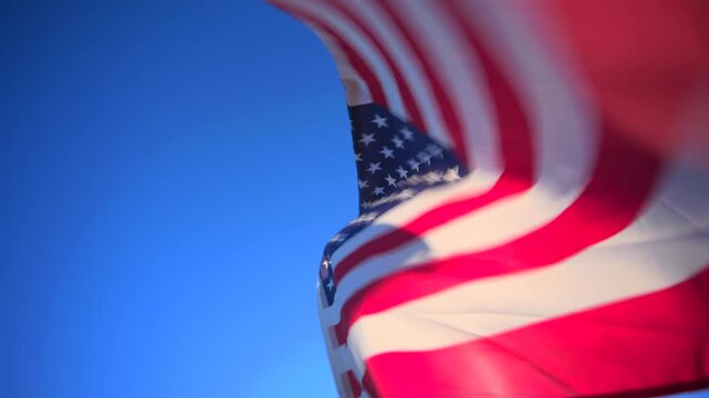 USA flag waves majestically against a vibrant clear blue sky. The iconic stars and stripes flutter in the wind showing red white and blue colors in soft focus with dynamic movement.
