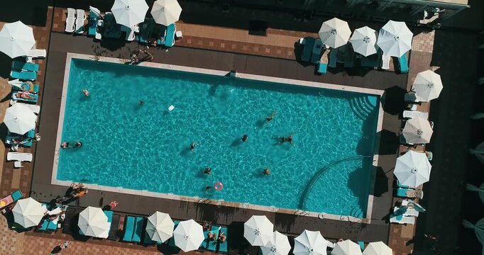 Aerial top down view of a rectangular blue swimming pool surrounded by white umbrellas and sun loungers at a resort