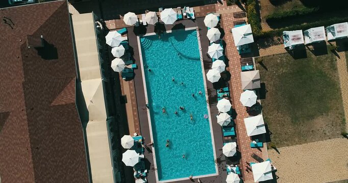 Aerial top down view of a rectangular blue swimming pool surrounded by white umbrellas and sun loungers at a resort