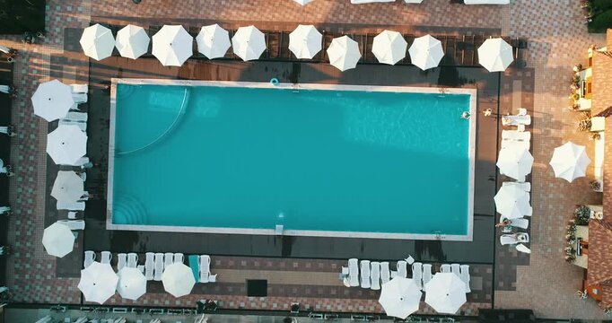 Aerial top down view of a rectangular blue swimming pool surrounded by white umbrellas and sun loungers at a resort